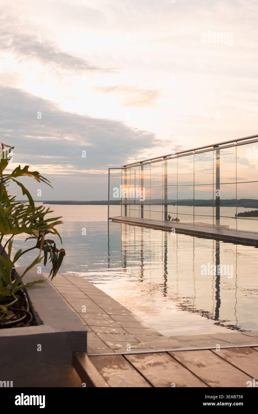 Les teintes douces du coucher de soleil brillent sur l'eau plate. La balustrade en verre reflète le reflet du ciel. La terrasse en bois s'étend vers la rive lointaine. Plante verte feuillue ajouter Banque D'Images
