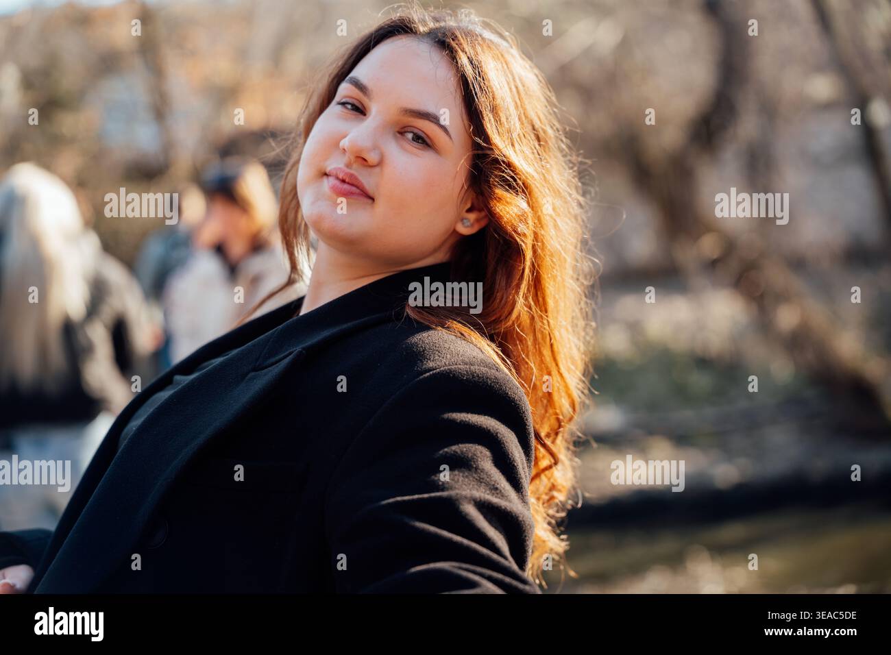 Jeune femme d'apparence caucasienne posant à l'extérieur dans le parc près de l'eau avec la lumière du soleil et un flou de fond doux. Portrait, style de vie, confiance. Banque D'Images