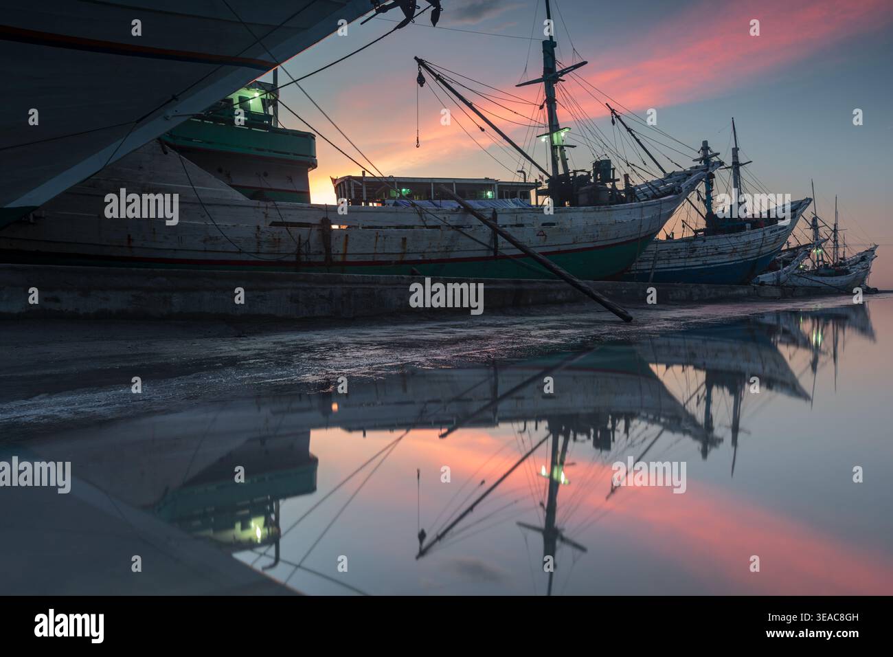 Les bateaux traditionnels Pinisi en bois amarrés au port de Sunda Kelapa à Jakarta, en Indonésie, se reflétant parfaitement dans l'eau calme lors d'un coucher de soleil vibrant Banque D'Images