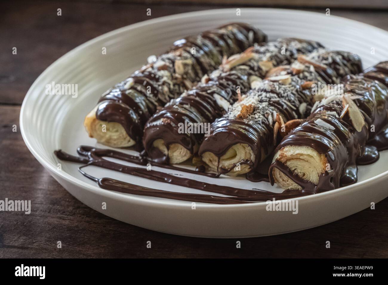 Gros plan photo de délicieuses crêpes à la sauce au chocolat et saupoudrées d'amandes sur une plaque blanche sur une planche de bois rustique. Crêpes de dessert fines, rouleau Banque D'Images