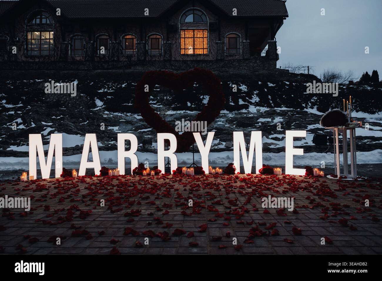 Scène de proposition de crépuscule avec lettres MARRY ME illuminées, pétales de rose dispersés sur la rive en bois, lanterne lumineuse, fenêtre de maison lointaine coulant chaud Banque D'Images