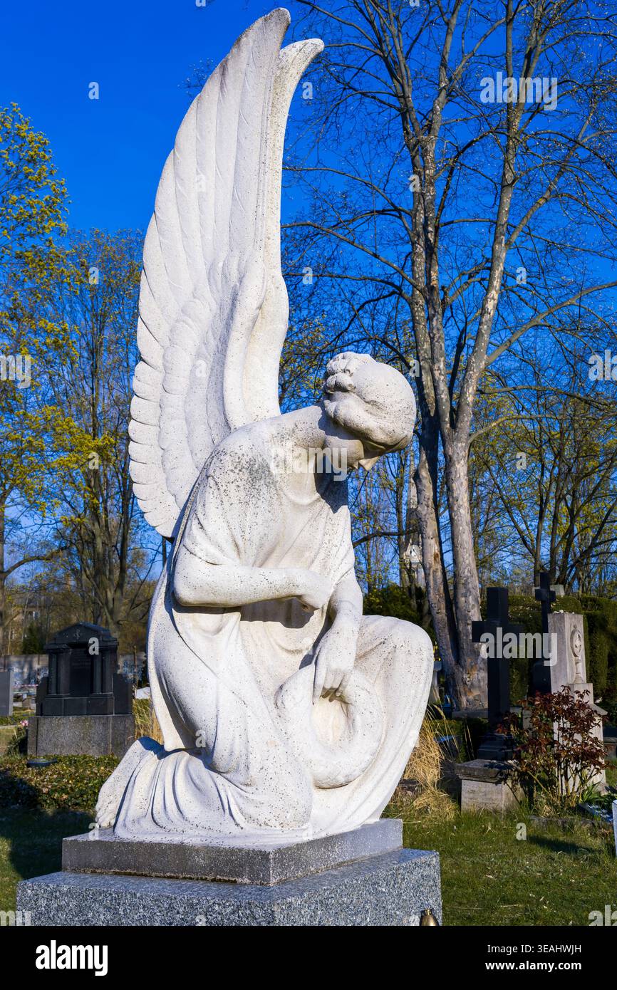 Sculpture d'ange en pierre dans une humble pose agenouillée sur une tombe de cimetière sous un ciel bleu clair, Berlin, Allemagne. Ange avec des ailes, Engelsskulptur. Banque D'Images