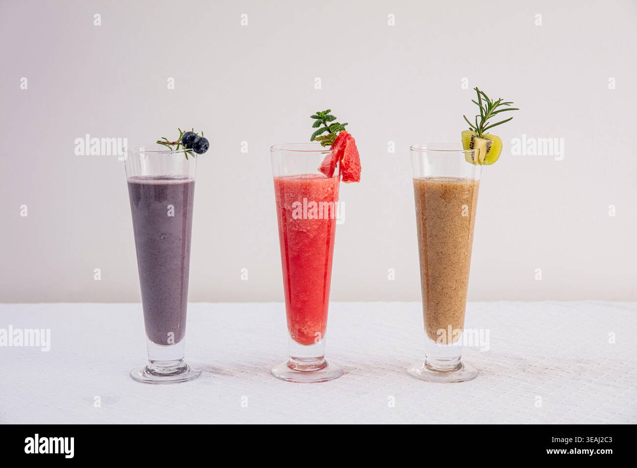 Trois grands verres de smoothies différents, chacun garni d'une garniture unique, reposent sur une nappe blanche dans un cadre lumineux et moderne. Banque D'Images