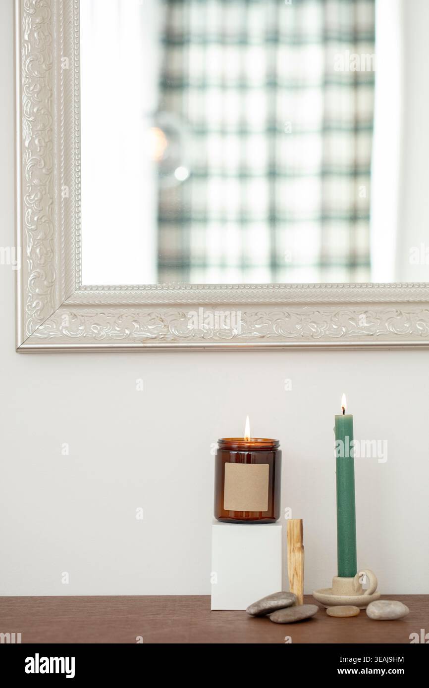 Une bougie allumée dans un bocal en verre se trouve sur une table en bois, entourée de cailloux et d'un bougeoir, devant un mur blanc et un miroir argenté. Banque D'Images