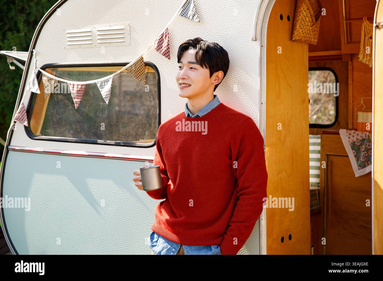 Un jeune homme se tient devant un camping-car vintage, tenant une tasse à café et souriant en profitant du soleil du matin. Banque D'Images