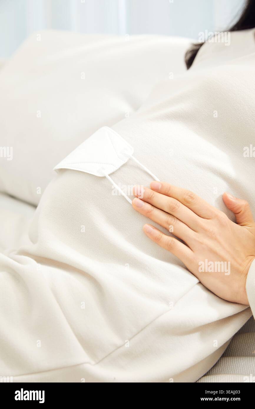 Une femme portant un masque facial est assise sur un canapé, berçant son ventre enceinte. Banque D'Images