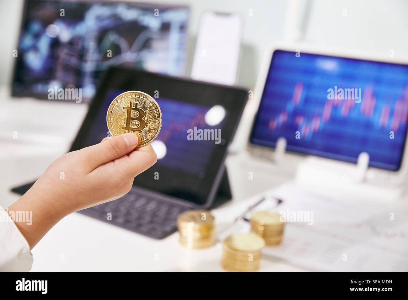 Une main tient une pièce Bitcoin devant un ordinateur portable et deux tablettes affichant des données financières. Banque D'Images