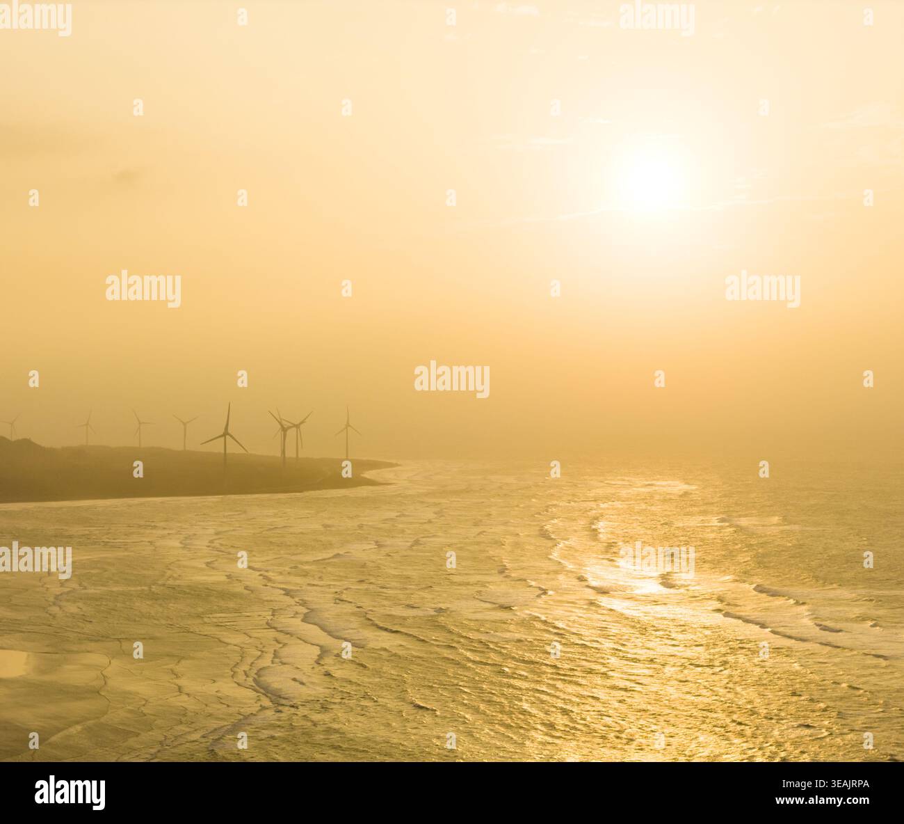 Vue aérienne d'éoliennes le long d'une côte brumeuse avec des vagues s'écrasant sur le rivage lors d'un coucher de soleil doré comté de Miaoli, Taiwan. Banque D'Images