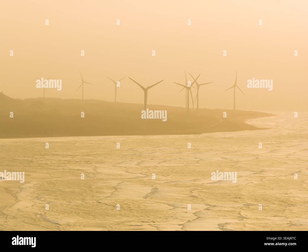 Vue aérienne d'éoliennes debout le long d'une côte brumeuse avec des vagues s'écrasant sur le rivage lors d'un coucher de soleil doré comté de Miaoli, Taiwan. Banque D'Images