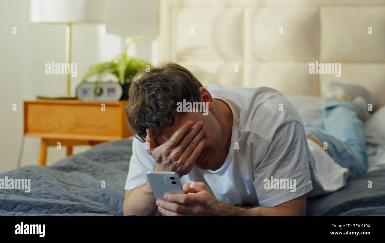 Homme caucasien inquiet allongé sur le lit à la maison tenant un téléphone portable saisissant la tête bouleversé homme choqué problème gars déçu par l'échec d'erreur de smartphone Banque D'Images