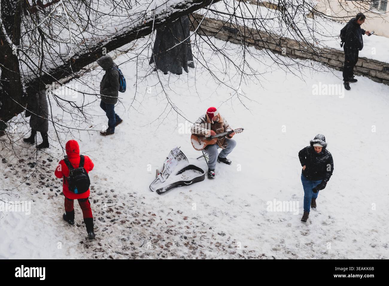 Tallinn, Estonie - 03 janvier 2025 : vue d'un guitariste solitaire faisant la sérénade des passants sur la place enneigée de l'hôtel de ville, une touche de manteaux rouges vibrants Banque D'Images