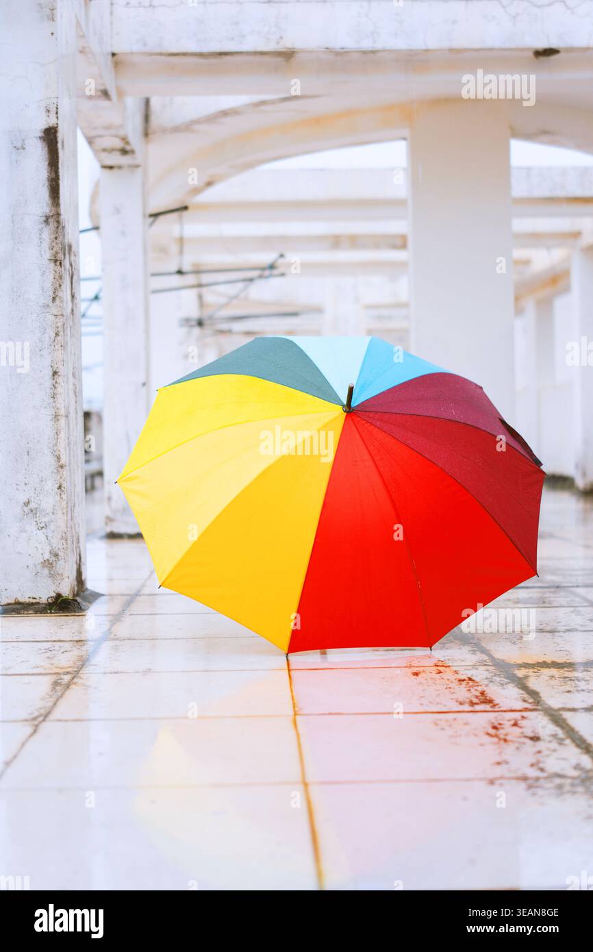 Parapluie arc-en-ciel coloré debout sur sol carrelé humide sous des arches blanches dans une zone urbaine abritée Banque D'Images