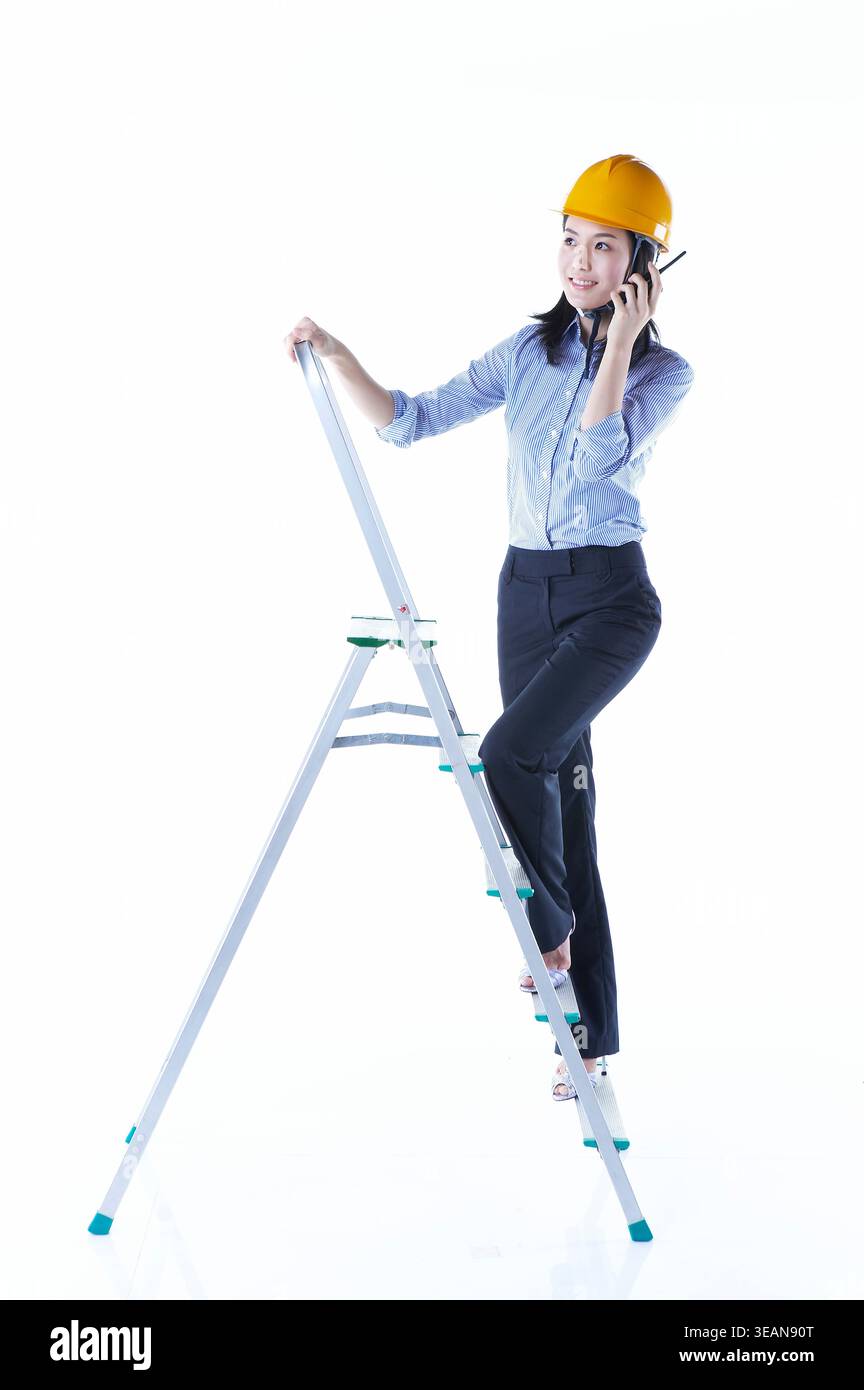 Une femme portant un casque de sécurité se tient debout sur une échelle tout en tenant un téléphone à son oreille. Banque D'Images