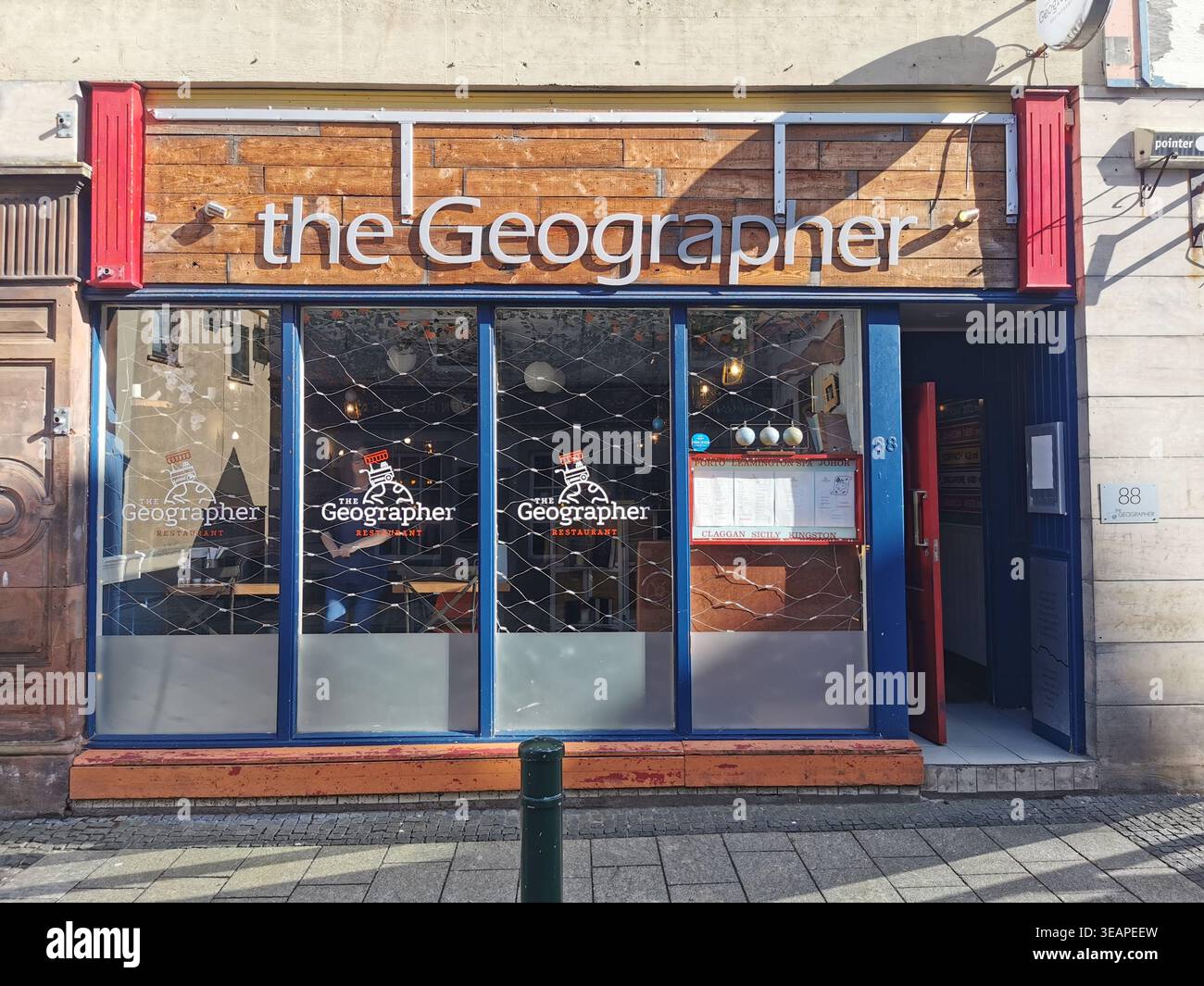 Devant le restaurant géographe sur Fort William High Street en plein soleil, sa signalétique se démarque dans les ombres de début de soirée. - Image de stock capturée avec un smartphone