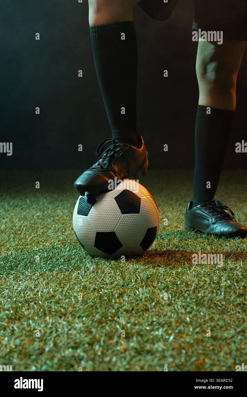 Femme afro-américaine équilibrant le crampon noir sur le ballon de football tout en portant des chaussettes hautes aux genoux sur le gazon. Athlète, chaussures, gros plan, dramatique, studio, brume, Banque D'Images
