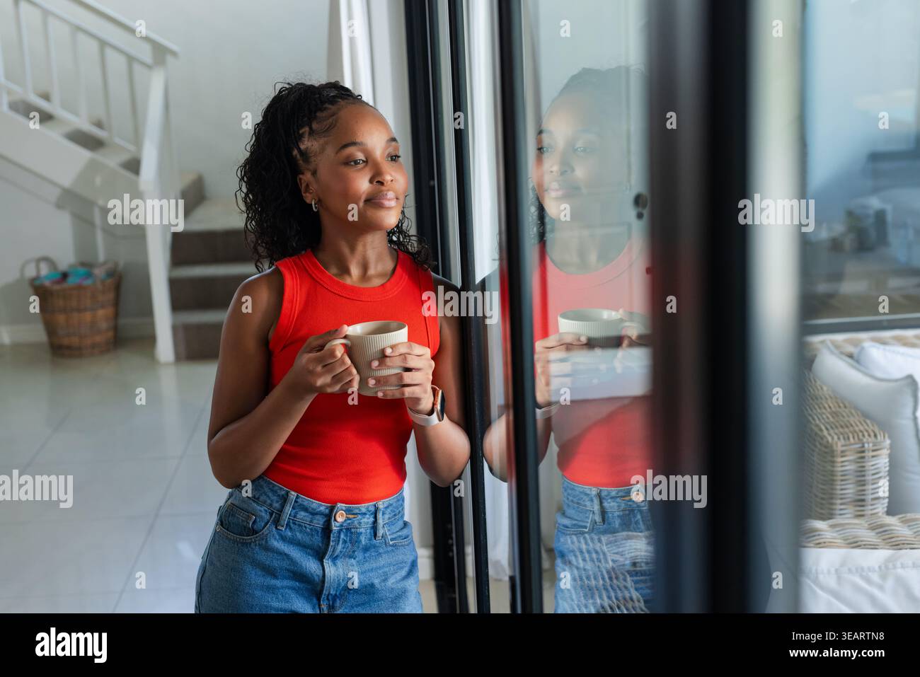 Femme africaine tenant une tasse, portant un haut rouge et un Jean à la maison, porte coulissante en verre reflet vu. Céramique, verre, lumière du jour, serein, moderne, minimaliste Banque D'Images
