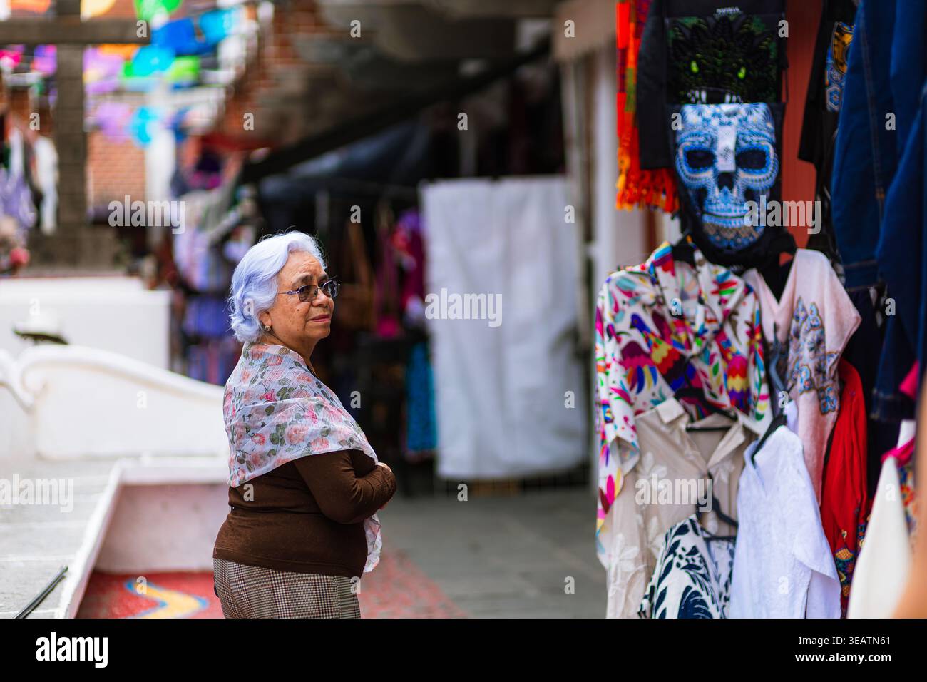 Une femme âgée regardant un étal présentant des souvenirs mexicains typiques, y compris des masques de crâne et des textiles, dans un marché traditionnel. Banque D'Images