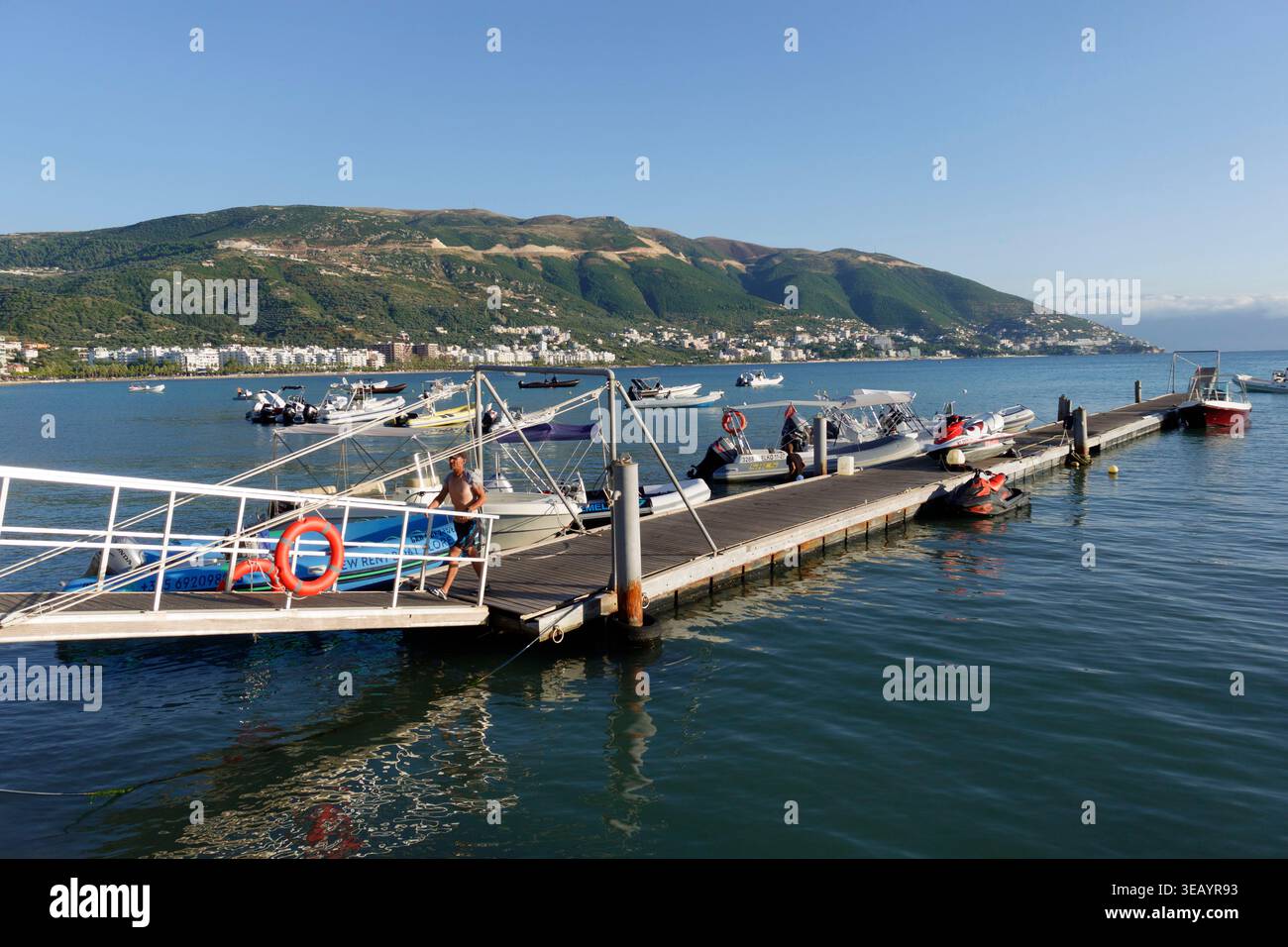 Bateaux et jet skis amarrés sur un quai flottant en bois avec une ville côtière et des montagnes en arrière-plan sous un ciel clair Banque D'Images