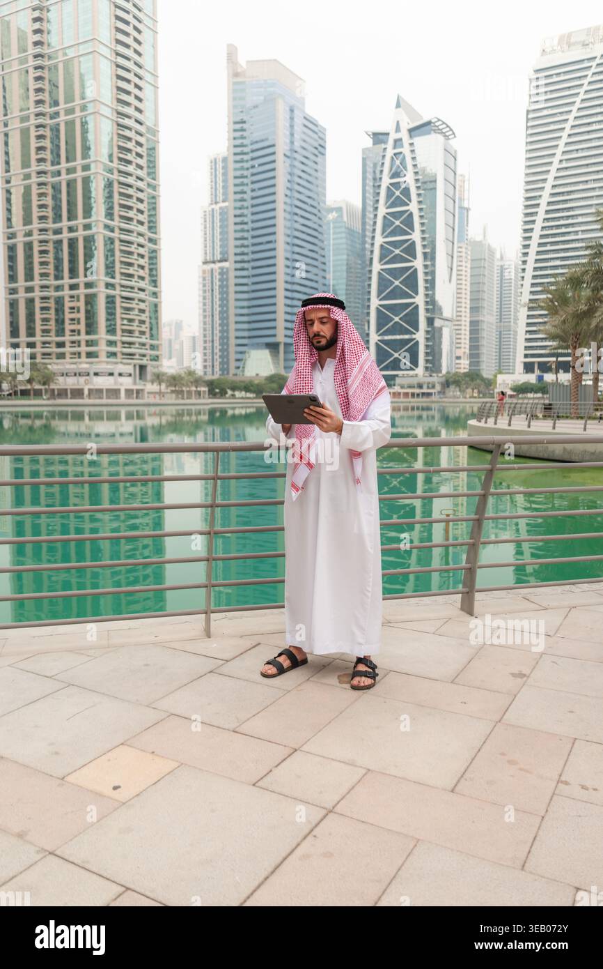 Homme d'affaires arabe en robe traditionnelle se tient près de l'eau dans une ville remplie de bâtiments modernes. Il tient un appareil tout en paraissant concentré. Banque D'Images