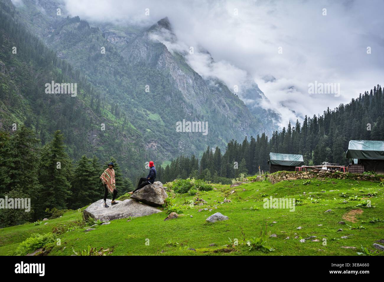 Sur un grand rocher, deux voyageurs s'arrêtent pour admirer le panorama époustouflant des montagnes embrassées par les nuages et la riche vallée verdoyante, profitant d'un moment de Banque D'Images
