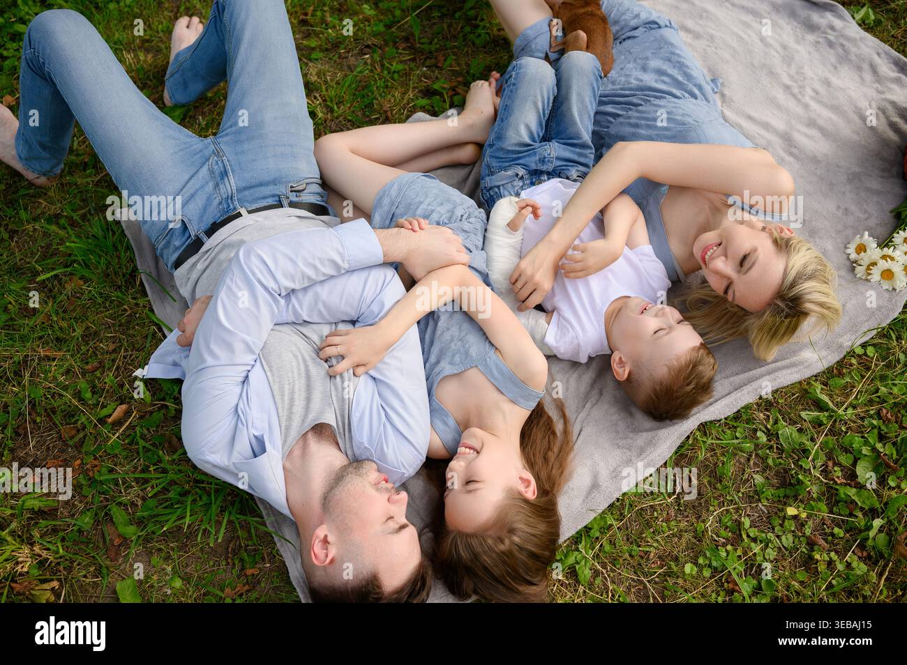 Vue de dessus de la famille heureuse couchée, câlin sur couverture dans le parc d'été. Concept d'environnement sûr, inclusion pour l'enfant avec moulage de bras et lien familial. Parents et enfants se relaxant sur l'herbe pendant le pique-nique Banque D'Images