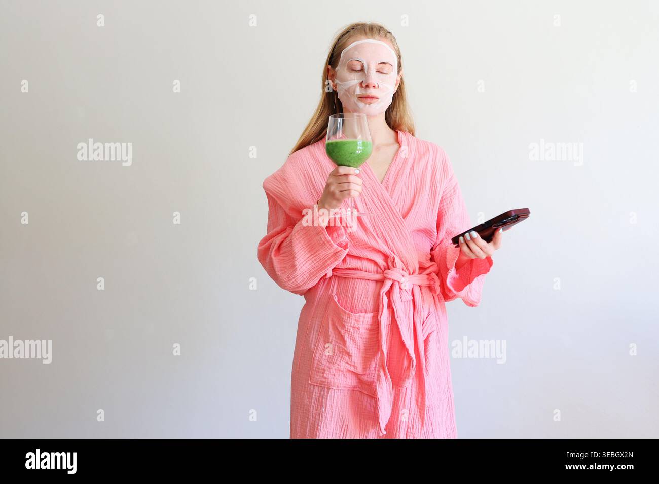 Femme dans le masque de feuille parlant sur smartphone avec smoothie vert, concept de soins personnels multitâches. Banque D'Images