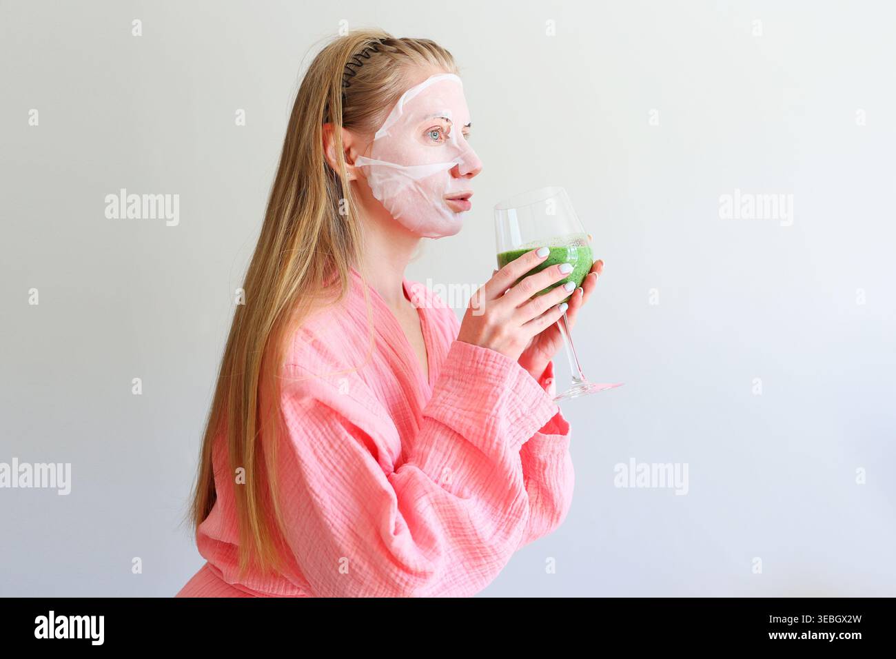 Femme détendue avec masque de feuille cosmétique et smoothie sain, rituel matinal d'auto-soin. Banque D'Images