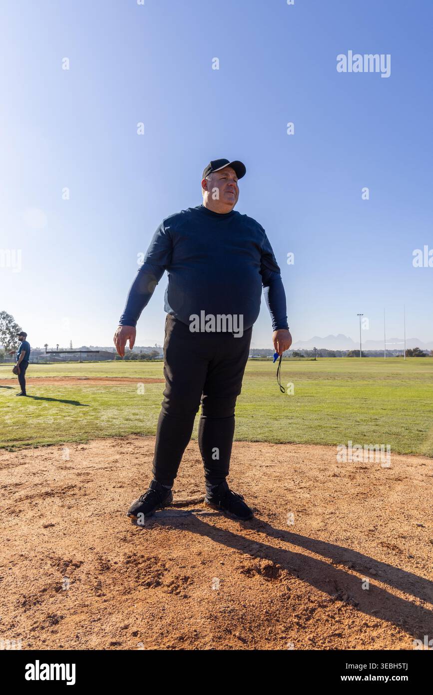 Homme senior debout sur un monticule de lanceur surélevé sur un diamant portant une casquette de baseball tenant des lunettes de soleil Banque D'Images