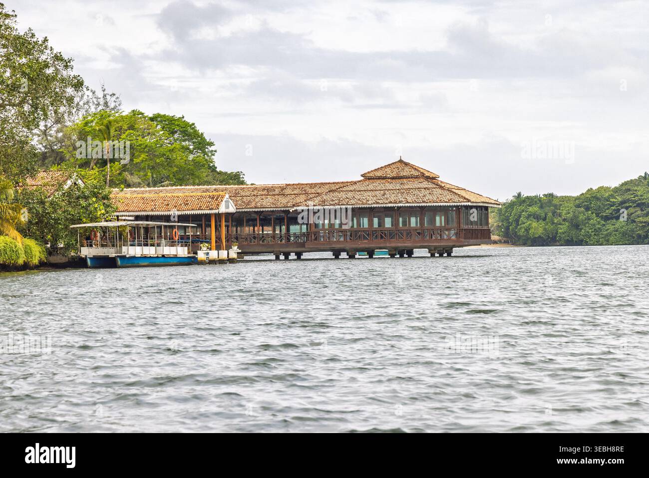 Restaurant Riverside sur pilotis avec un toit de chaume traditionnel, entouré d'une verdure tropicale luxuriante et d'une eau calme sous un ciel nuageux. Sri Lanka isl Banque D'Images