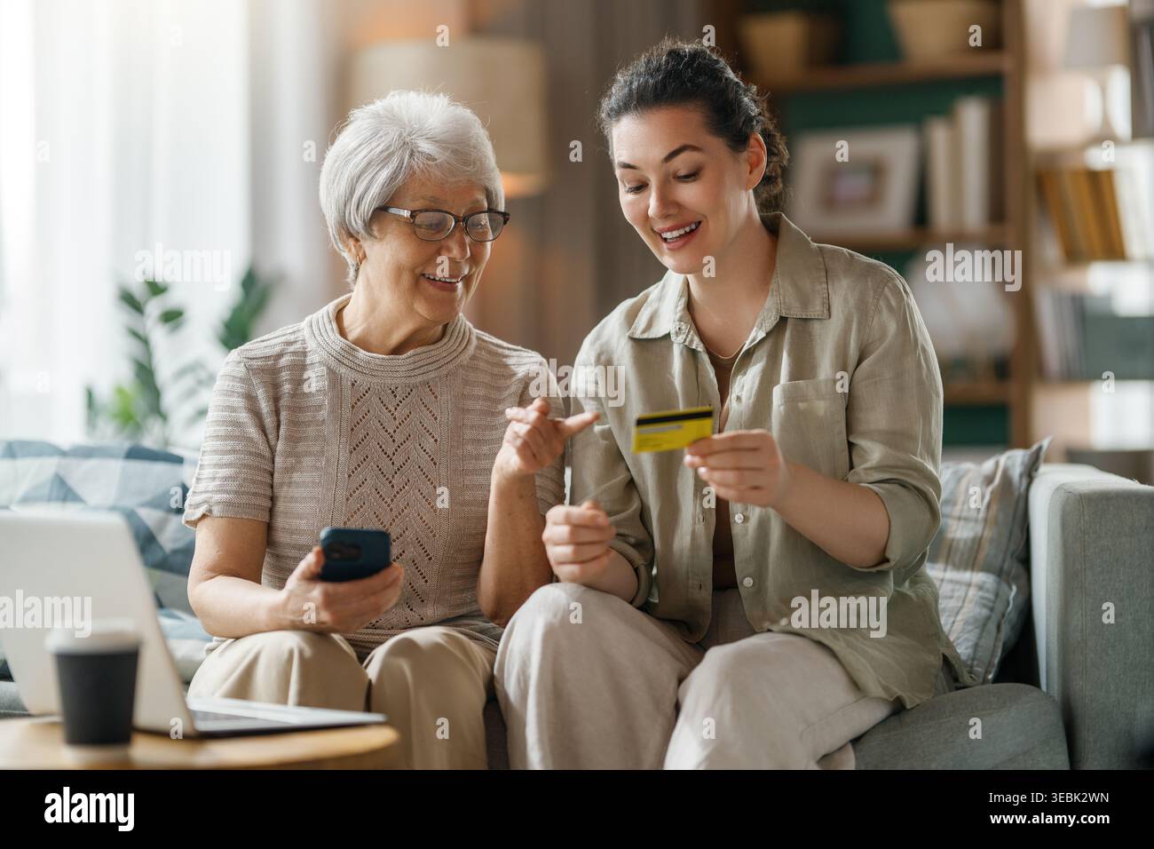 Joyeuse femme âgée et sa fille adulte faisant un achat en ligne ensemble à la maison. La paire est assise confortablement sur un canapé confortable, partageant un moment de Banque D'Images