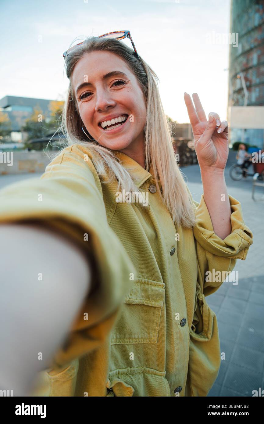 Jeune femme joyeuse prenant heureux un selfie tout en montrant ludique signe de paix avec ses doigts en plein soleil, rayonnant de joie et de positivité avec Banque D'Images