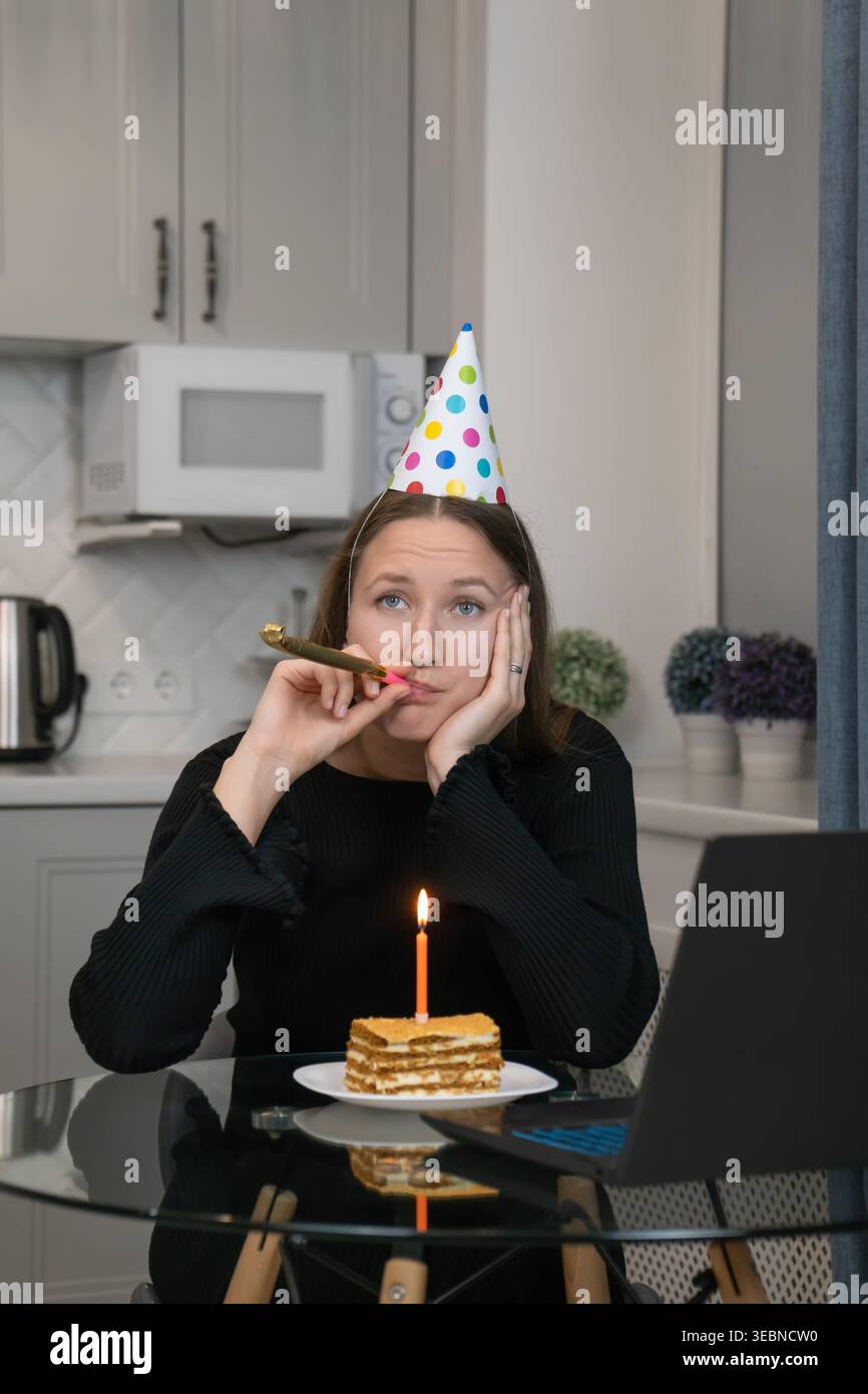 Femme triste portant chapeau de fête soufflant corne de fête et célébrant l'anniversaire en ligne à l'aide d'un ordinateur portable, assis à la table de cuisine avec morceau de gâteau et bougie allumée Banque D'Images