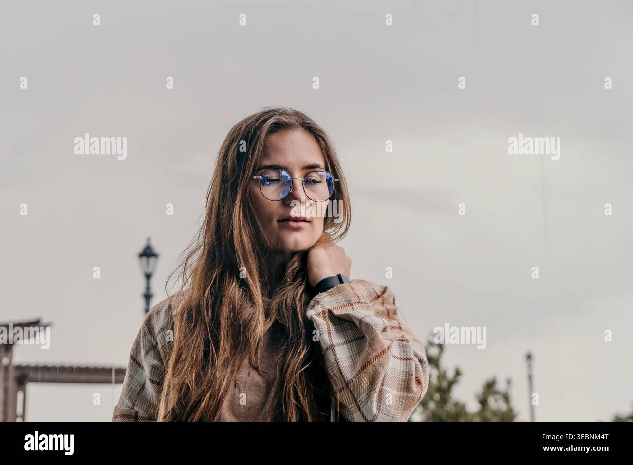 Femme lunettes dehors pensive jeune femme portant des lunettes rondes et une chemise à carreaux contre un ciel nuageux avec espace de copie Banque D'Images