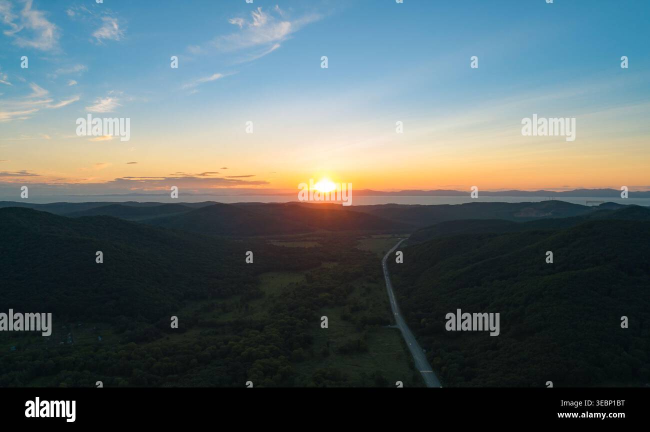La vue aérienne capture un coucher de soleil vibrant descend sur des montagnes verdoyantes et une route sinueuse dans une vallée paisible au crépuscule Banque D'Images