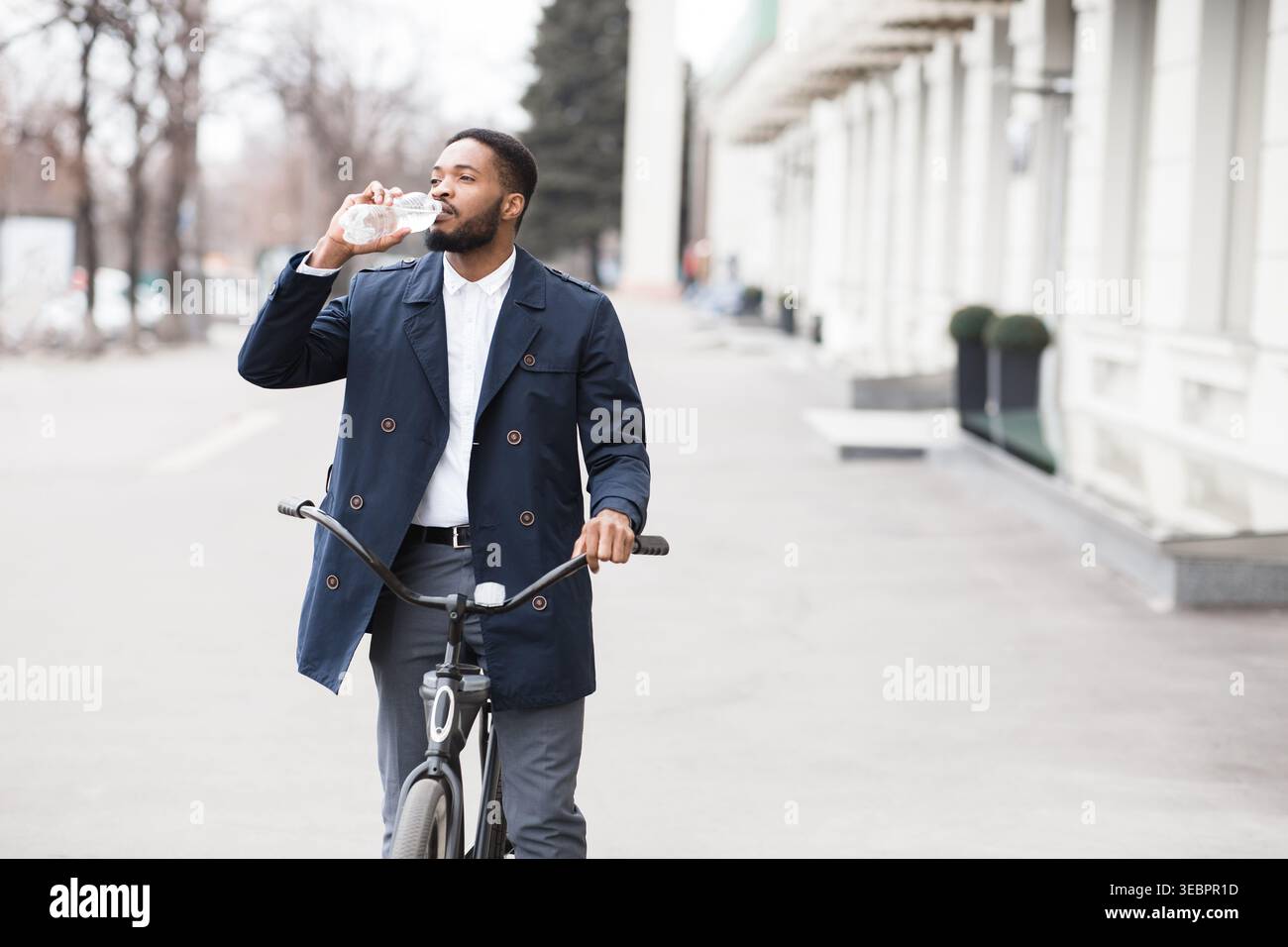Homme portant un manteau boit de l'eau tout en faisant du vélo dans un environnement urbain un jour ensoleillé Banque D'Images