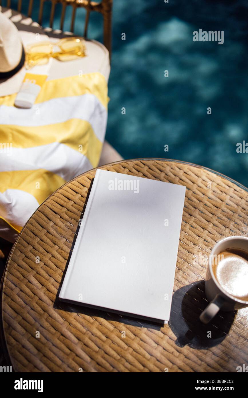 Cahier vierge repose sur une table en osier à côté d'une tasse de café et d'une serviette rayée jaune, avec une piscine visible en arrière-plan Banque D'Images