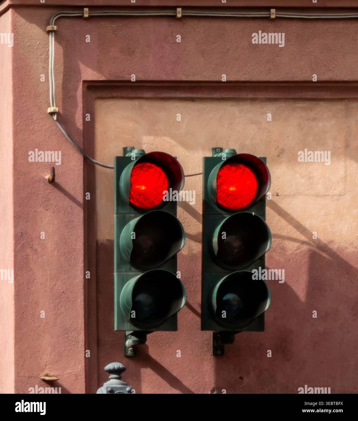 Deux feux rouges montés sur un mur de bâtiment texturé en terre cuite. Les câbles électriques et les ombres nettes sont visibles en plein jour. Banque D'Images