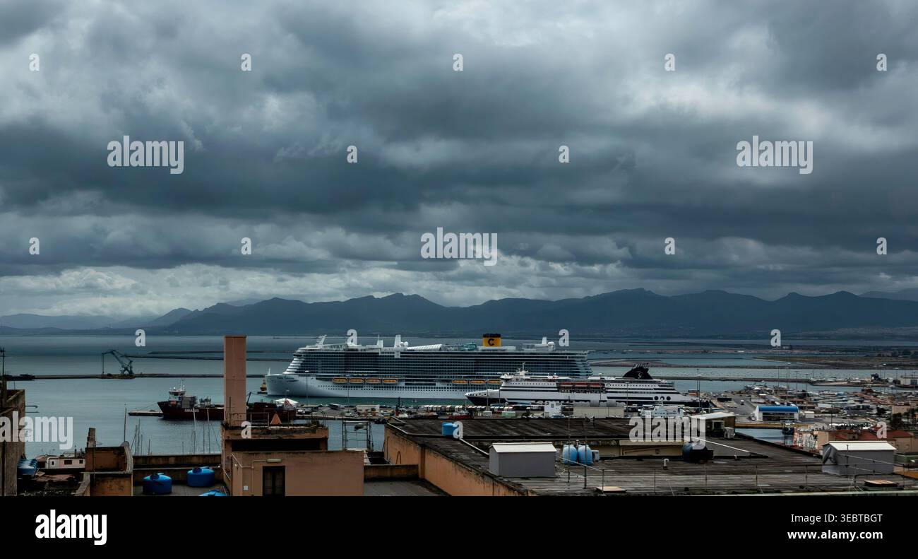 Un grand bateau de croisière et ferry accosté dans le port de Cagliari. Des nuages sombres et orageux surplombent le port et les montagnes lointaines de Sardaigne. Banque D'Images