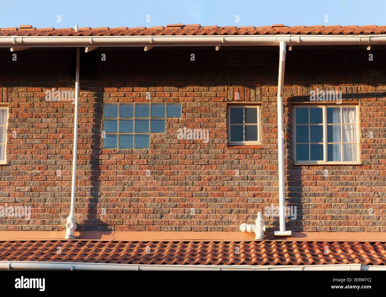 détail d'une maison en briques brunes avec des fenêtres en blocs de verre, des fenêtres traditionnelles et des tuyaux de descente blancs sous un toit de tuiles sous la lumière du jour claire. Banque D'Images