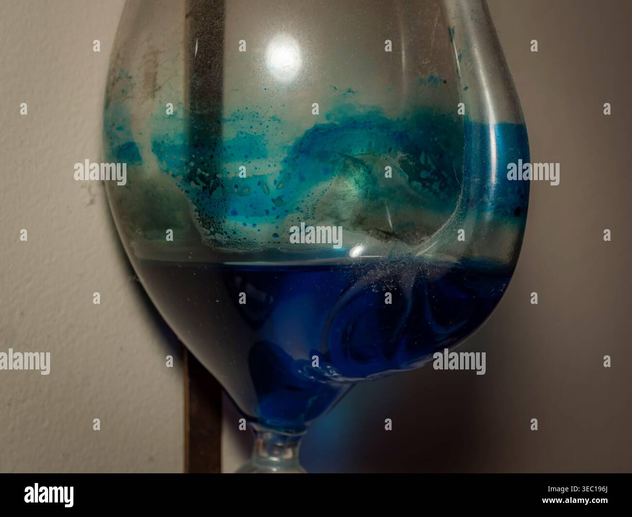 Vue macro d'un sablier en verre sale montrant des couches de liquide bleu et sarcelle avec des résidus de moisissure ou de poussière contre un mur Banque D'Images