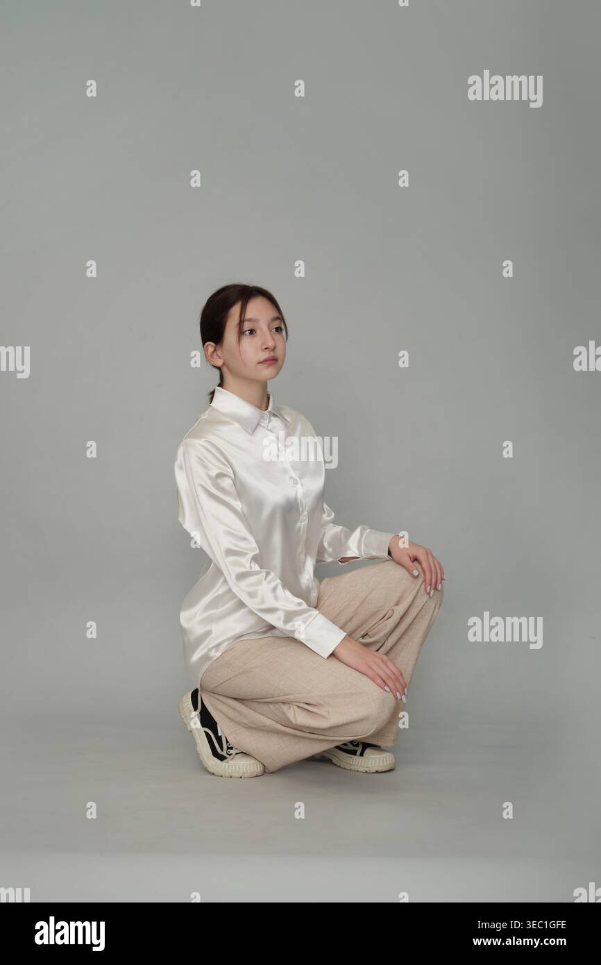 Une jeune femme dans une blouse blanche satinée et un pantalon beige s'accroupit soigneusement sur un fond neutre. Cette image souligne l'élégance de Banque D'Images