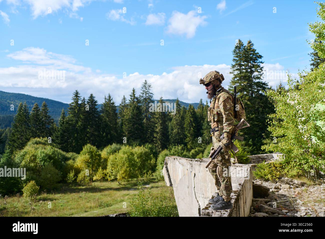 Soldat camouflé debout sur le mur en ruine surplombant le paysage de montagne boisé avec équipement tactique Banque D'Images