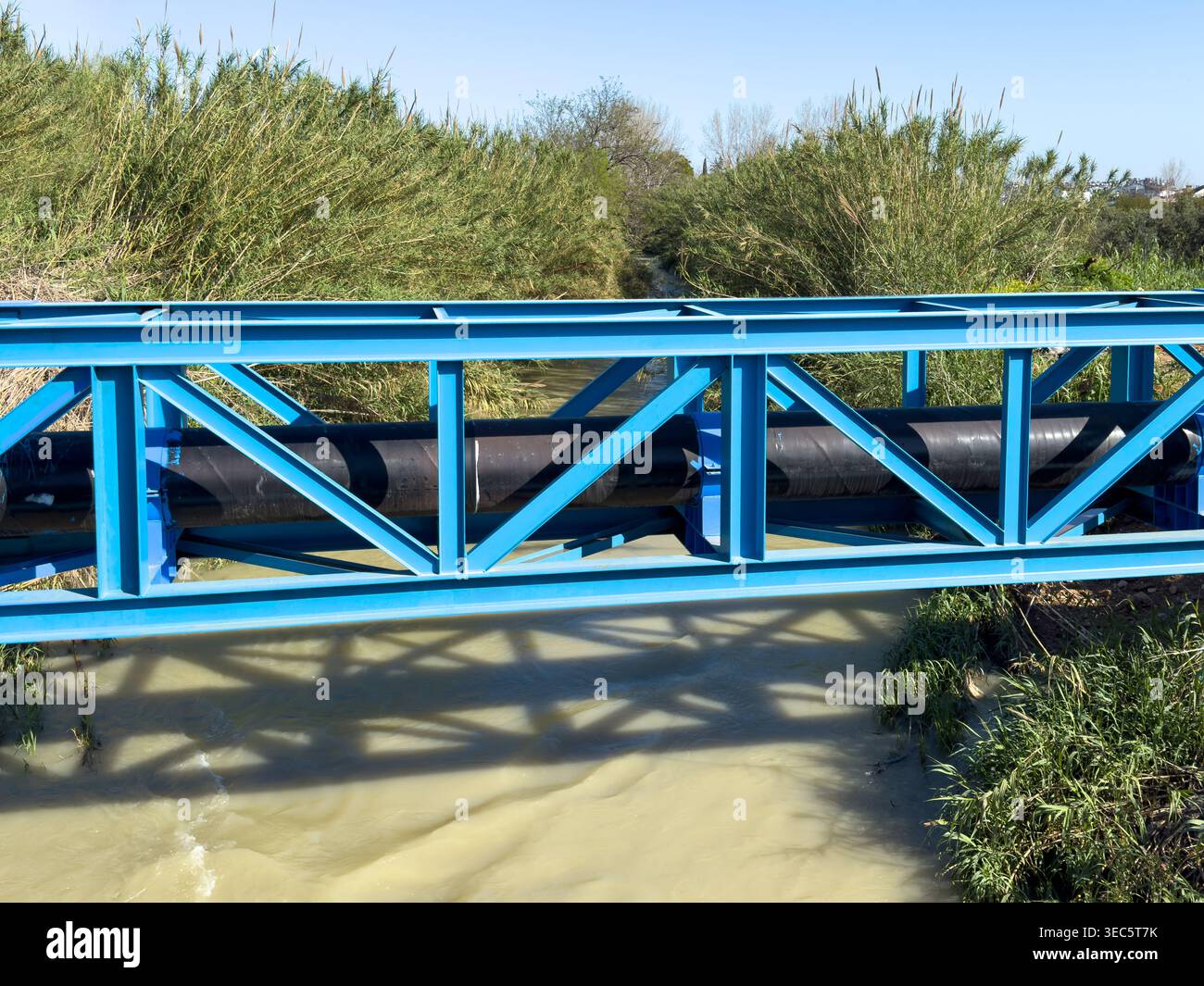 Pont en treillis d'acier bleu soutenant un grand pipeline industriel au-dessus d'une rivière qui coule Banque D'Images