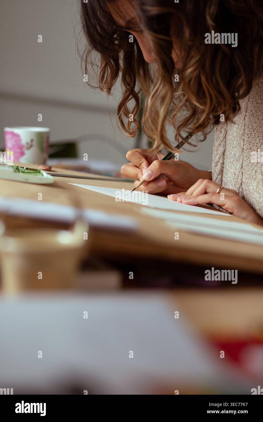 Une femme est assise à un bureau concentré sur le dessin avec un crayon taillé. Elle travaille avec soin, entourée de matériel de croquis et d'une tasse à proximité. Le shi léger Banque D'Images