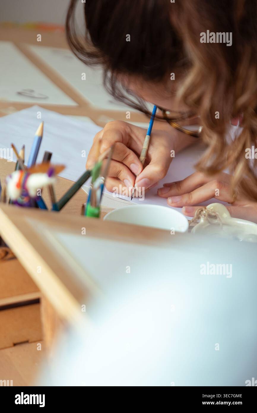Une femme est assise à un bureau, peignant soigneusement des lettres avec un pinceau fin. Elle se concentre intensément sur sa tâche, entourée d'outils et de matériaux. Le voyant c Banque D'Images