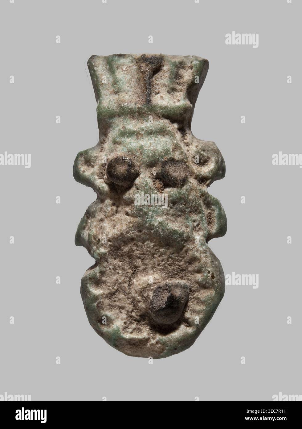 Amulette — tuile simple face avec une représentation schématique en relief de Bes : une tête avec une couronne à plumes et un bouclier avec un phallus . Yeux et phallus appliqués avec glaçage noir. Amulette, plâtre/carreaux de céramique. Période tardive–période ptolémaïque, VIIe–Ier siècle av. J.-C., Égypte. Hauteur 2,7 cm ; largeur 1,3 cm., un objet de forme irrégulière avec une surface rugueuse et piquée dans les tons de brun, de brun et de vert est montré sur un fond gris neutre ; la forme a un sommet plus large qui s'effile légèrement puis se bombe en saillies arrondies le long des côtés, avec une paire de bosses arrondies surélevées près du milieu supérieur et Banque D'Images