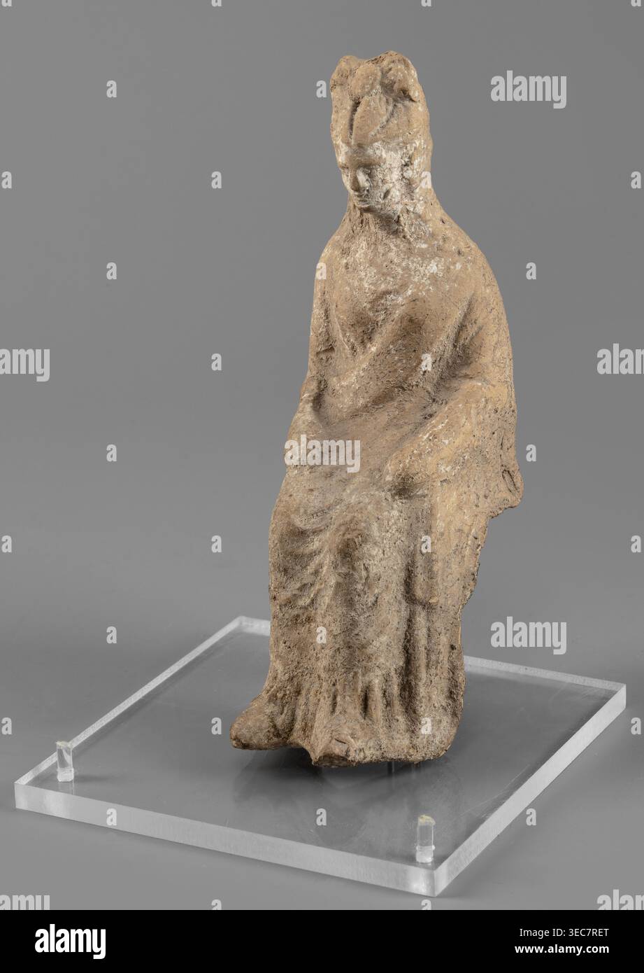 Figurine féminine en position assise, légèrement penchée en avant. Le dos de la figurine n'est pas artistiquement développé. La femme est vêtue d'une robe et d'un manteau révélant l'épaule droite et tombant presque aux chevilles. Ses mains sont placées sur ses genoux. Ses cheveux sont attachés dans un chignon haut. Traces de peinture blanche sur la tête de la figurine. IVe siècle av. J.-C., Italie du Sud. Figurine en terre cuite, hauteur 15,4 cm, largeur 5 cm, profondeur 7,3 cm, poids 0,17 kg., une figurine assise faite de matériau texturé rugueux repose sur une base rectangulaire claire, avec une palette discrète de brun et beige sur la figure et un gris Banque D'Images