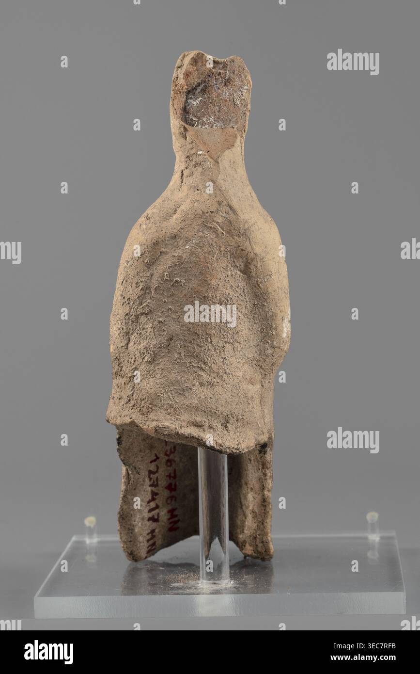 Figurine féminine, figure féminine en position assise, légèrement penchée en avant. Le dos de la figurine n'est pas artistiquement développé. La femme est vêtue d'une robe et d'un manteau révélant l'épaule droite et tombant presque aux chevilles. Ses mains sont placées sur ses genoux. Ses cheveux sont attachés dans un chignon haut. Traces de peinture blanche sur la tête de la figurine. IVe siècle av. J.-C., Italie du Sud. Figurine en terre cuite, hauteur 15,4 cm, largeur 5 cm, profondeur 7,3 cm, poids 0,17 kg., une forme sculpturale usée ressemblant à une figure humaine simplifiée se tient sur une base rectangulaire claire avec un support vertical clair ; la figure Banque D'Images
