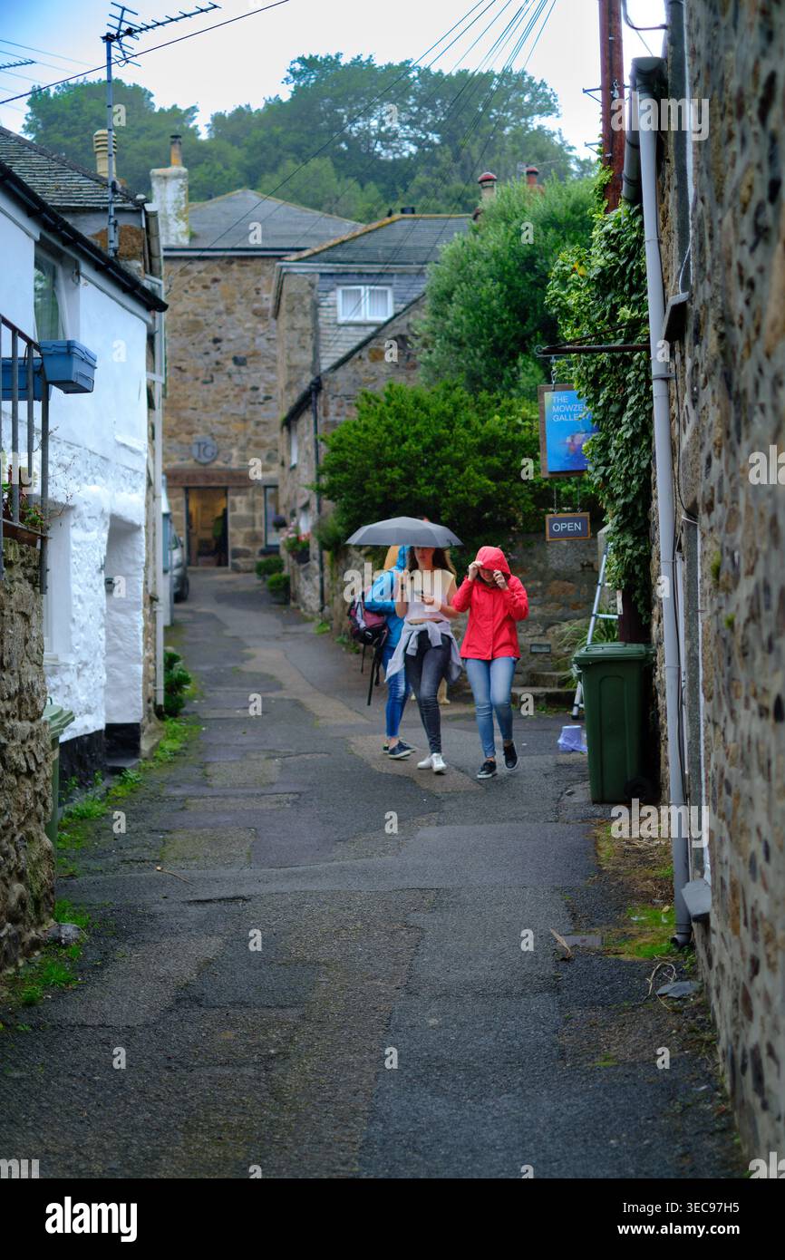 Les touristes marchent sous un parapluie le long d'une étroite ruelle pluvieuse devant des chalets en pierre et une galerie d'art dans le village côtier de Mousehole, Cornouailles, Englan Banque D'Images
