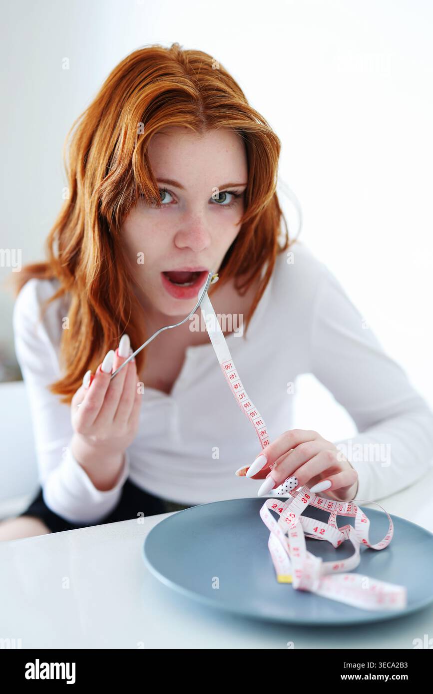 Femme rousse prétendant manger du ruban à mesurer, anorexie, régime strict et métaphore de contrôle des calories. Banque D'Images